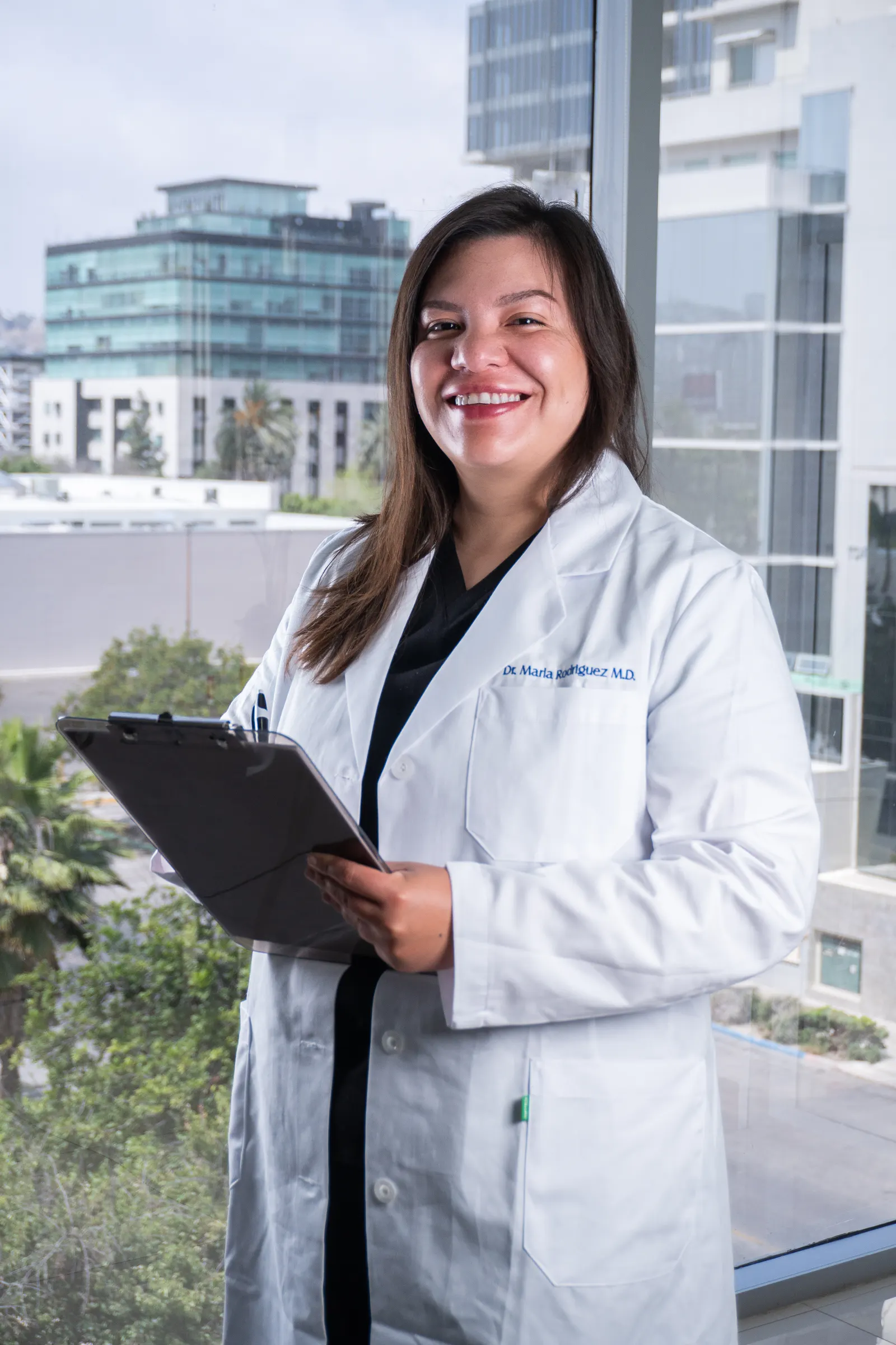 Dra. Marla Rodriguez holding a clipboard at chest level, white lab coat with 'Dr. Marla Rodriguez M.D.' embroidered, reviewing patient notes