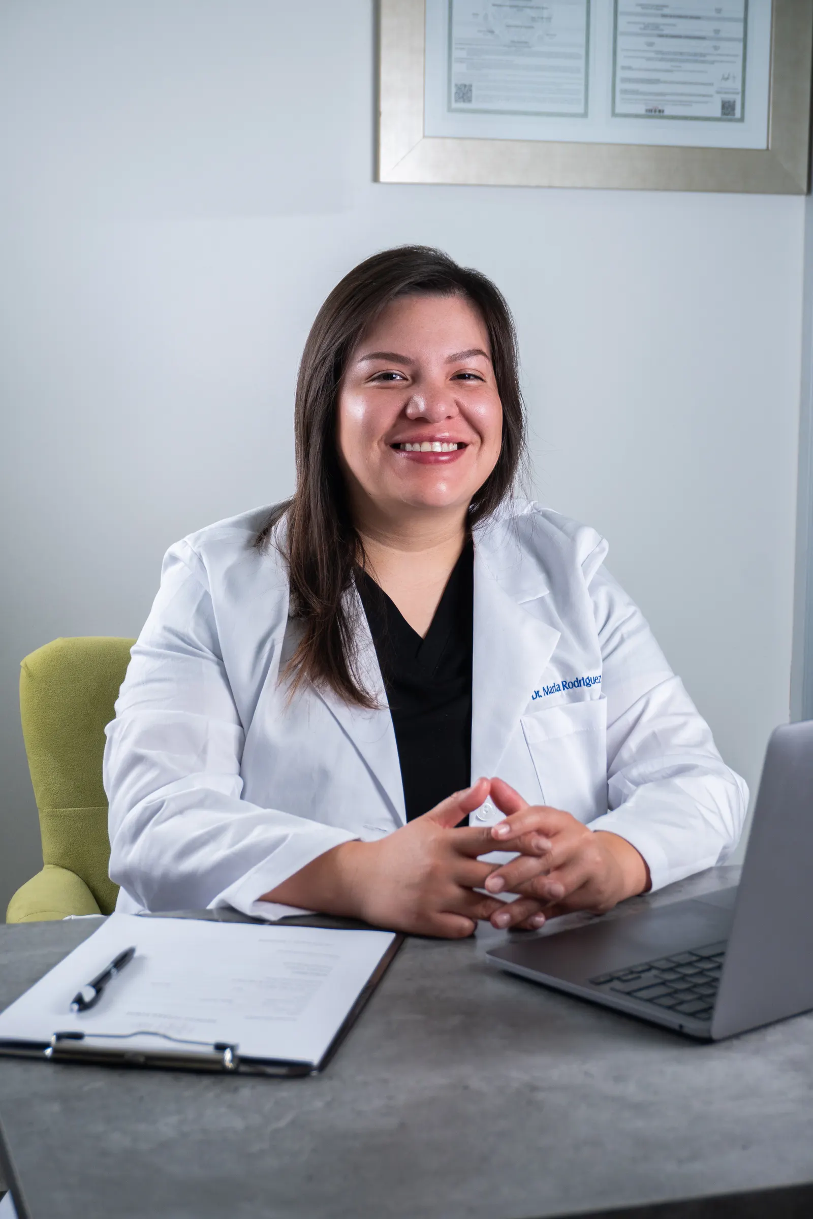 Dra. Marla Rodriguez at her desk with a MacBook and patient charts, the framed medical-degree credential visible on the wall behind her