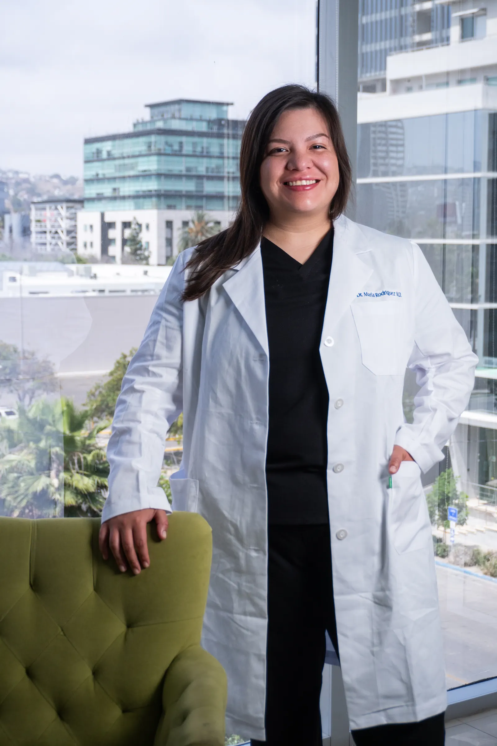 Dra. Marla Rodriguez in a white lab coat over black scrubs, hand resting on a green velvet chair in the consultation suite