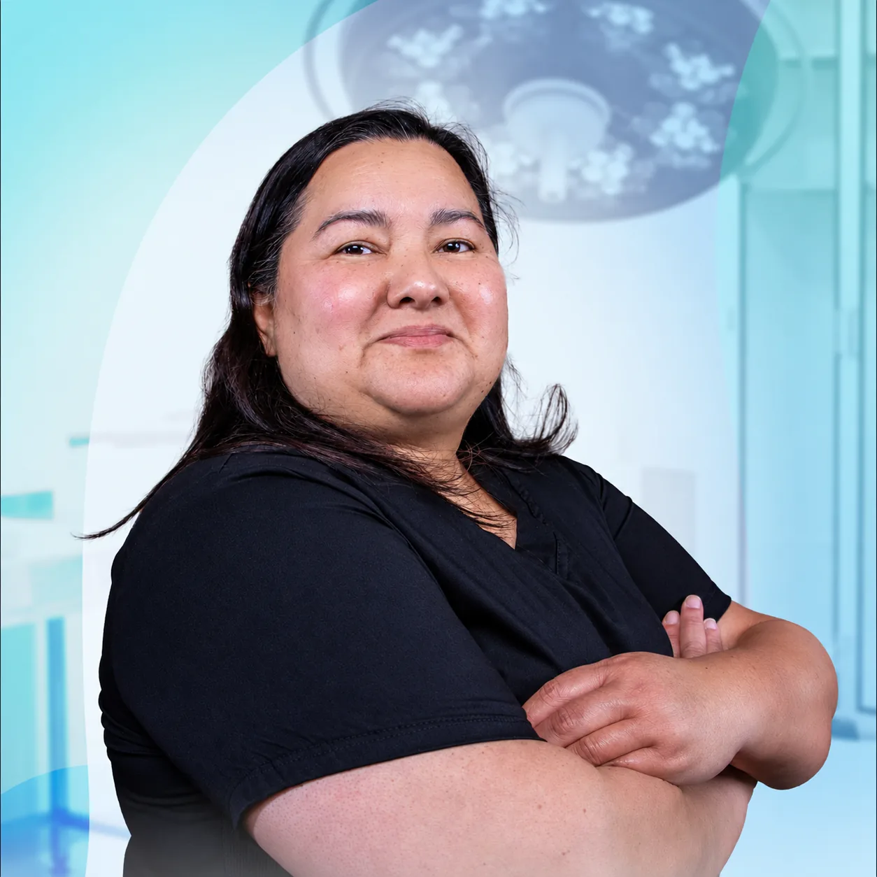 Dra. Silvia Castillo, board-certified plastic surgeon, in black scrubs with arms crossed beneath the operating-room lights