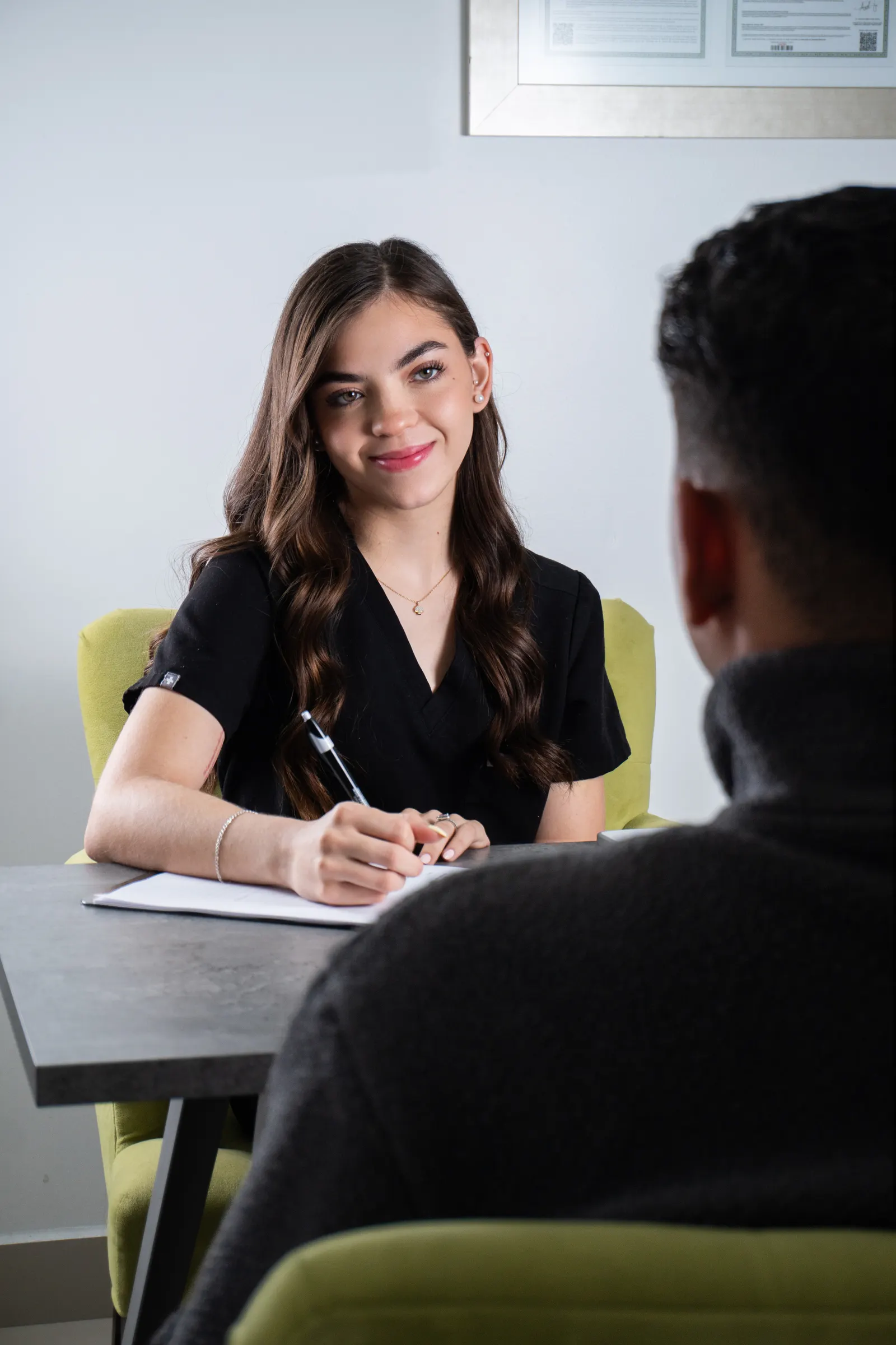 Natalia Gonzalez at her desk taking notes during a new-patient consultation, framed credential plaque visible on the wall behind her