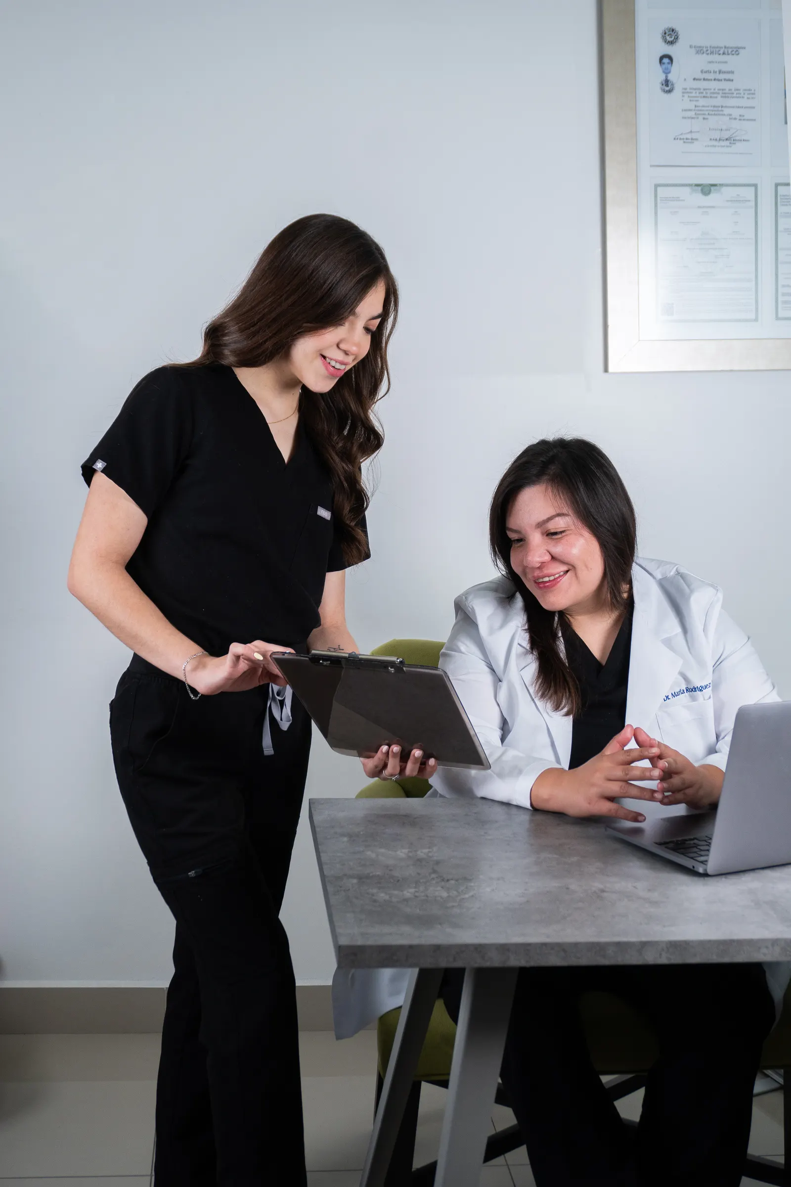 Dra. Marla Rodriguez seated at her desk reviewing a clipboard with Natalia Gonzalez standing beside her, the framed M.D. credential visible on the wall behind